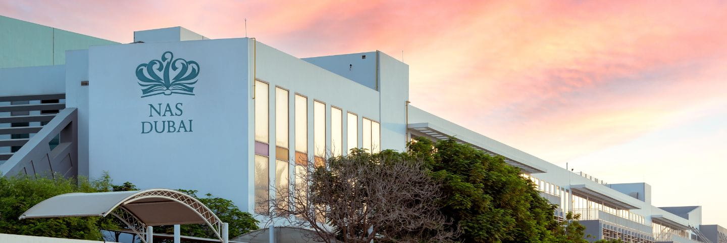 Modern exterior of NAS Dubai school building at sunset with the school logo visible, large windows, surrounding greenery, and a dusky sky.
