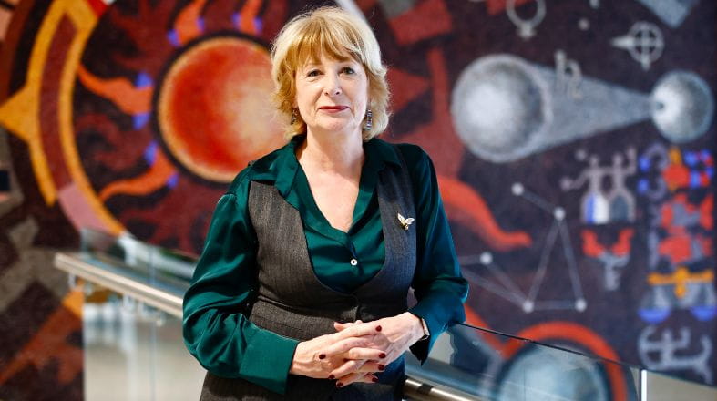 A person stands indoors beside a glass railing, wearing a dark vest over a green shirt, with a vibrant, abstract mural featuring geometric shapes and bold colors in the background.