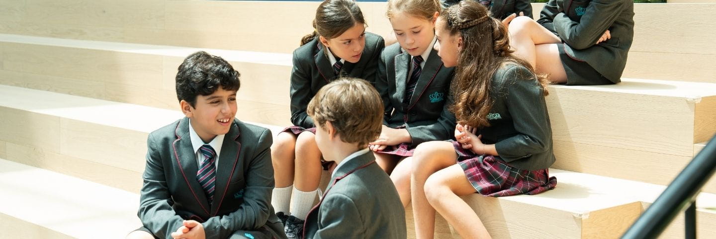 Several students in matching school uniforms sit together on wide wooden steps, engaged in conversation in a bright, modern indoor area.