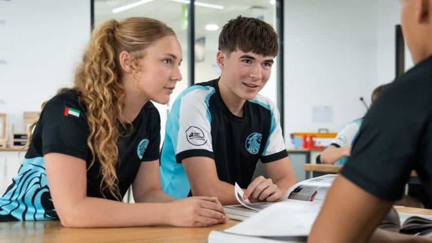 Two NAS Dubai students in school sports uniforms sitting at a classroom table, engaged in discussion and working from open books.
