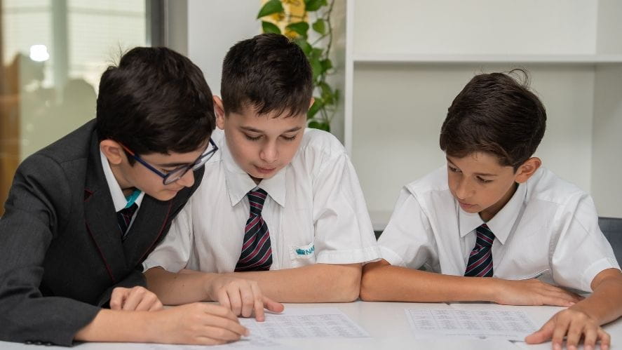A group of three NAS Dubai students wearing school uniforms sit at a table, closely examining printed worksheets and working together in a classroom setting.