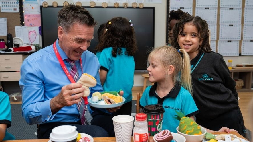 The NAS Dubai principal sitting with young students in a classroom, participating in a learning activity with play food and classroom materials on the table.