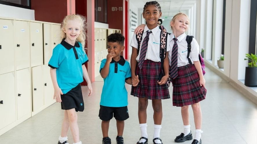 A group of four NAS Dubai students stand together in a bright school corridor lined with lockers. Some students wear turquoise PE uniforms while others wear the school’s plaid uniform with white shirts and ties. They appear to be interacting and holding hands as they prepare for their school day.