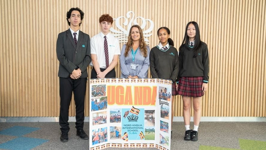 A group of five individuals standing indoors behind a large display board labeled “UGANDA,” which includes photos and information about a school‑support project in Uganda. The individuals are dressed in school uniforms, and the board features images related to the fundraising and awareness initiative.