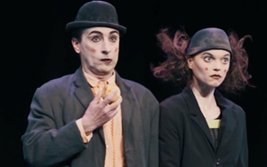 Two actors performing, a man in a bowler hat and a woman with her hair blown back are schocked.